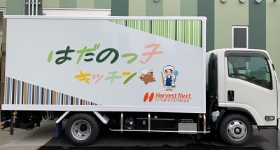 優しい色合いの文字で「はだのっこキッチン」と書かれ、調理師とボンチーヌが描かれた給食運搬用トラックの写真