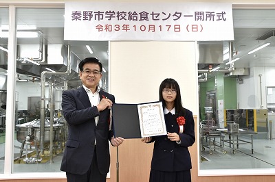 新しい学校給食センターの調理設備を背景に、スーツ姿の市長と制服姿の三嶽 愛望さんが表彰状を持って並んで立ち記念撮影している写真