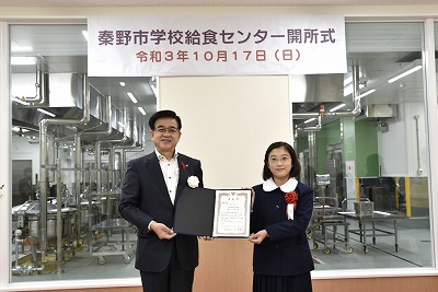 新しい学校給食センターの調理設備を背景に、スーツ姿の市長と制服姿の林 和泉さんが表彰状を持って並んで立ち記念撮影している写真