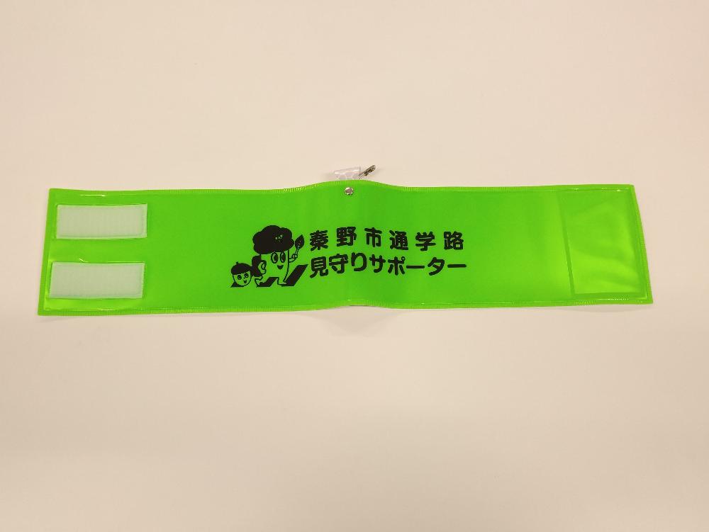 白い背景の上に広げられた蛍光グリーンの腕章で、「秦野市通学路見守りサポーター」という黒文字と、横断歩道をランドセルを背負ったもりりんが渡っているイラストが描かれた写真