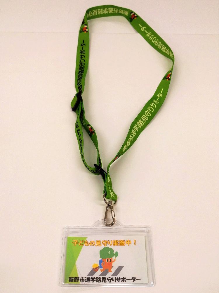 ネックストラップ付きのIDカードケースで、緑色のストラップには「秦野市通学路見守りサポーター」の文字とイラストが繰り返し印刷され、ケース内のカードには「子どもの見守り実施中！」の文字と横断歩道をランドセルを背負ったもりりんが渡っているイラストが描かれている写真
