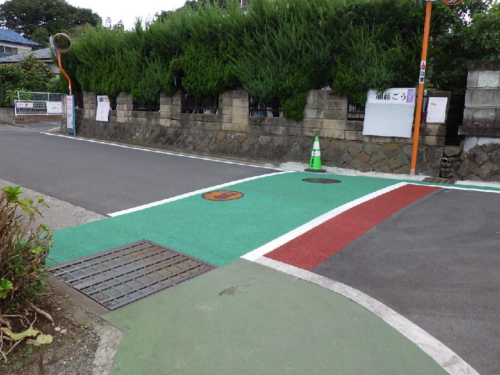 住宅地の道路の交差点の角で、鮮やかな緑色と赤色のカラー舗装が施された歩行者への注意喚起エリアが路面標示されている道路の写真