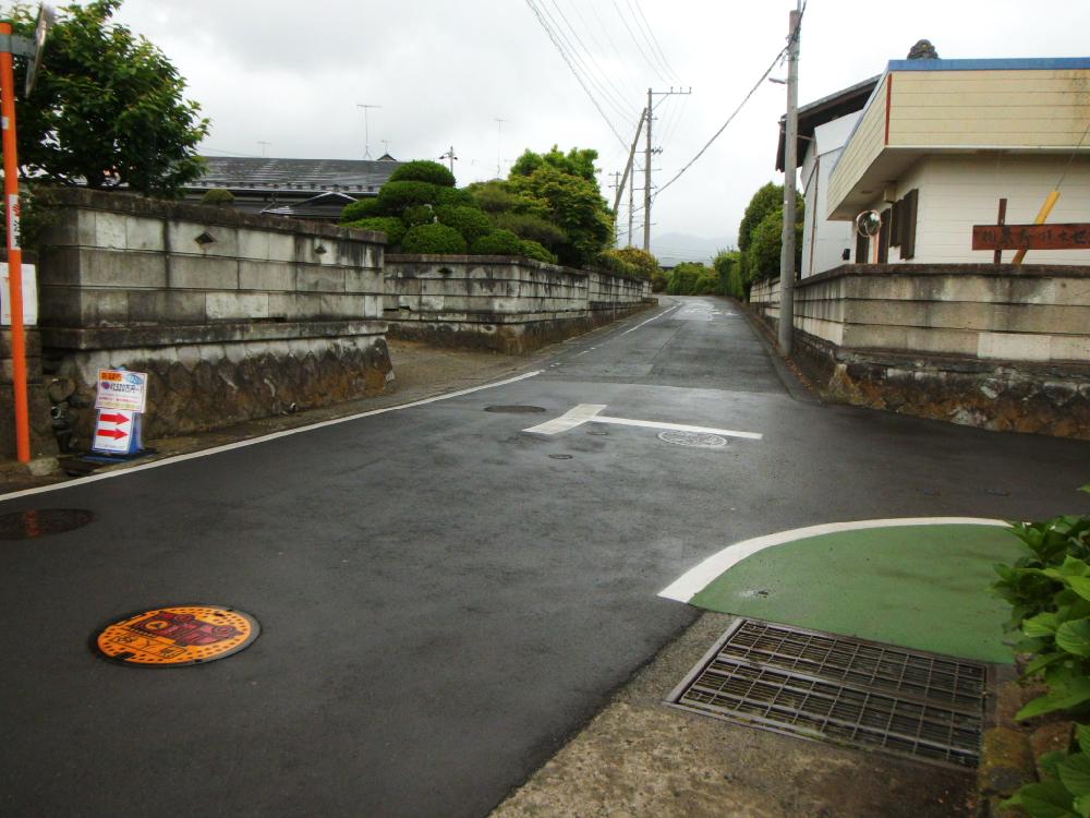 両側は石積みとブロック塀が並ぶ住宅街の道路に白いT字が路面標示されている道路の写真