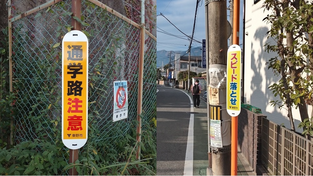 金網フェンスに、「通学路注意」と太字で書かれた縦長の黄色い標識が取り付けられ右側のオレンジ色の電柱のようなポールに、「スピード落とせ」と太字で書かれた縦長の黄色い標識が取り付けられている写真