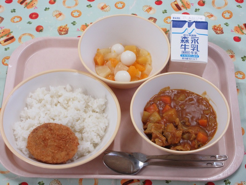 ピンクのトレーに給食が右上から反時計回りで牛乳、白玉フルーツポンチ、ご飯とメンチカツ、カレーライスと並べられている写真