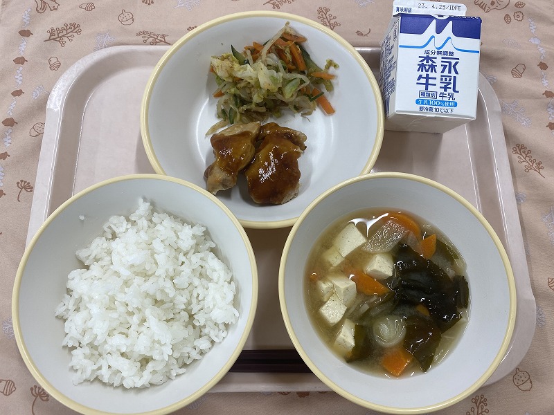 ピンクのトレーに給食が右上から反時計回りで牛乳、鶏もも肉の甘酒照り焼きと野菜のおかか和え、ご飯、味噌汁と並べられている写真