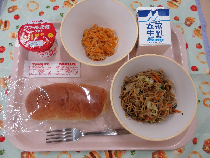 ピンクのトレーに給食が右上から反時計回りで牛乳、にんじんしりしり、ヨーグルト、チーズパン、ソース焼きそばと並べられている写真