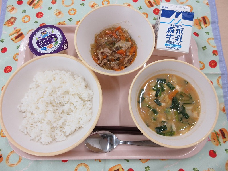 ピンクのトレーに給食が右上から反時計回りで牛乳、チャプチェ、ブルーベリーゼリー、ご飯、味噌チゲと並べられている写真