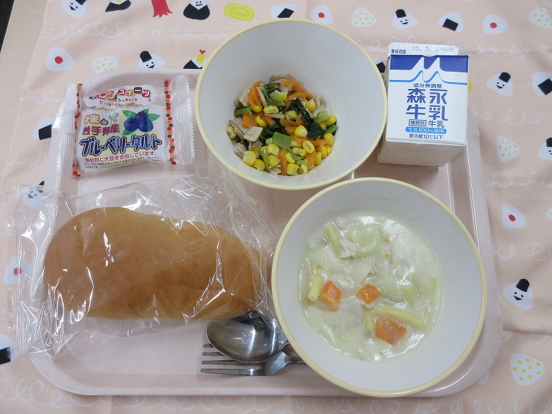 ピンクのトレーに給食が右上から反時計回りで牛乳、コーンソテー、ブルーベリータルト、コッペパン、マカロニのクリーム煮と並べられている写真