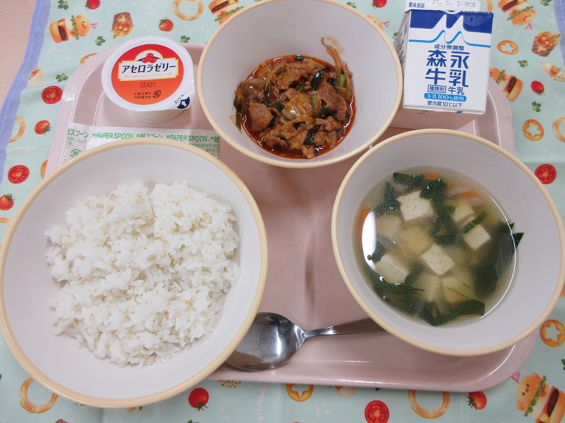 ピンクのトレーに給食が右上から反時計回りで牛乳、豚キムチ丼の具、アセロラゼリー、もち麦入りご飯、豆腐スープと並べられている写真