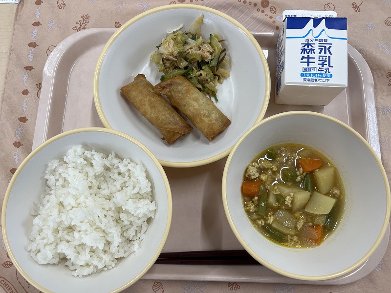ピンクのトレーに給食が右上から反時計回りで牛乳、春巻きとささみとキャベツの昆布和え、ご飯、じゃがいものカレー風味煮と並べられている写真