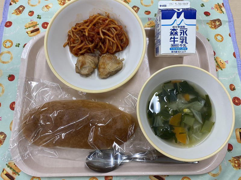 ピンクのトレーに給食が右上から反時計回りで牛乳、鶏肉のハーブ焼きとミートソーススパゲティ、米粉コッペパン、野菜スープと並べられている写真