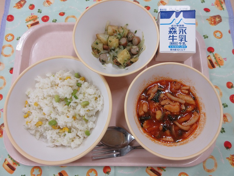 ピンクのトレーに給食が右上から反時計回りで牛乳、古谷さん家のジャーマンポテト、枝豆とコーンのピラフ、チキンと野菜のトマトソース煮込みと並べられている写真