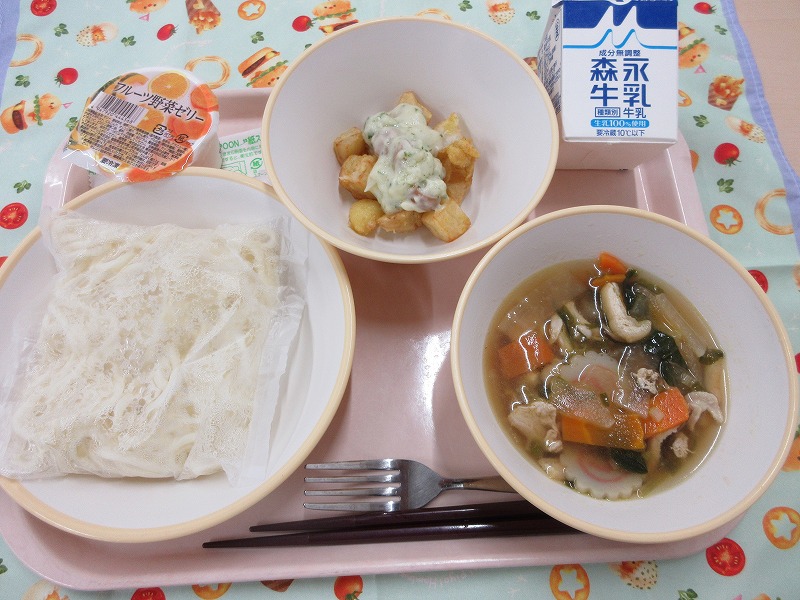 ピンクのトレーに給食が右上から反時計回りで牛乳、ポテトのチーズソース、フルーツ野菜ゼリー、ソフトめん、五目うどんと並べられている写真