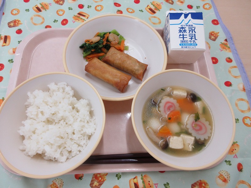 ピンクのトレーに給食が右上から反時計回りで牛乳、春巻きと青菜炒め、ごはん、白玉スープと並べられている写真