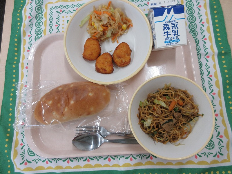 ピンクのトレーに給食が右上から反時計回りで牛乳、はりはりサラダとチキンナゲット、玄米パン、ソース焼きそばと並べられている写真