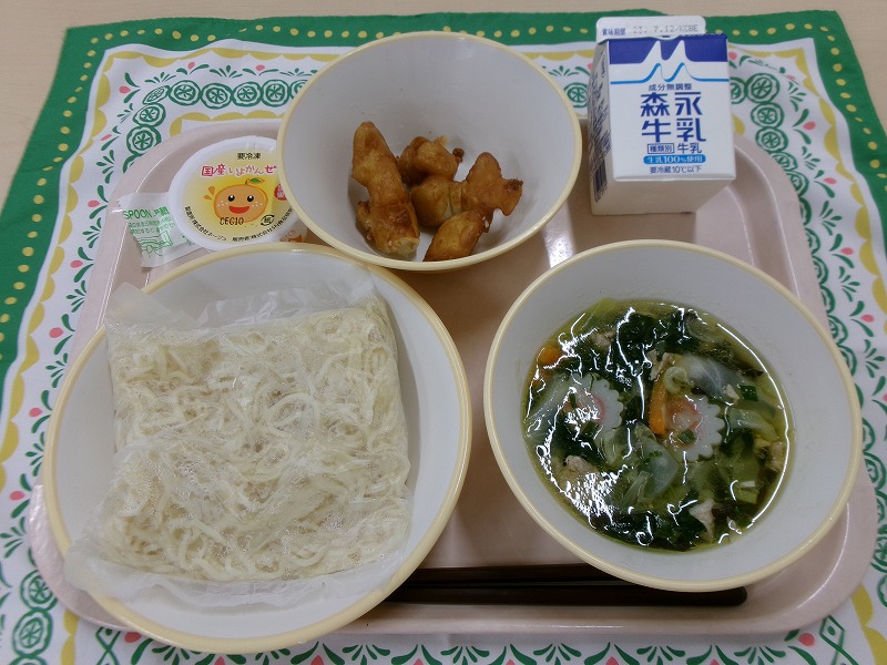 ピンクのトレーに給食が右上から反時計回りで牛乳、あげいも、いよかんゼリー、麺、五目塩ラーメンと並べられている写真