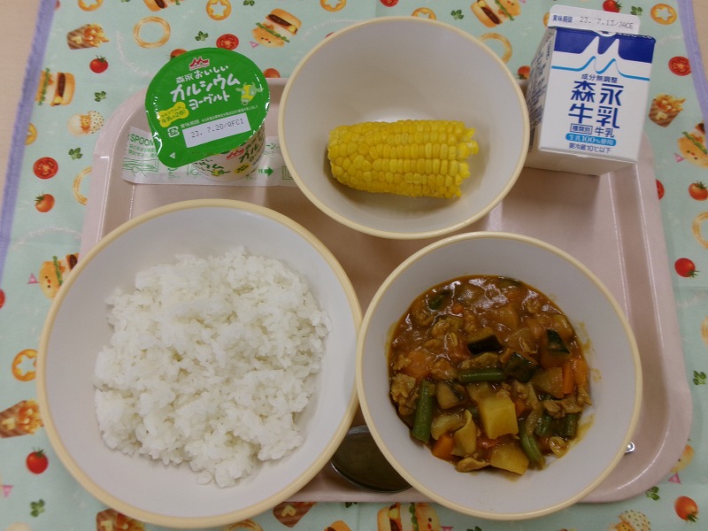 ピンクのトレーに給食が右上から反時計回りで牛乳、とうもろこし、ヨーグルト、ごはん、夏野菜のカレーと並べられている写真