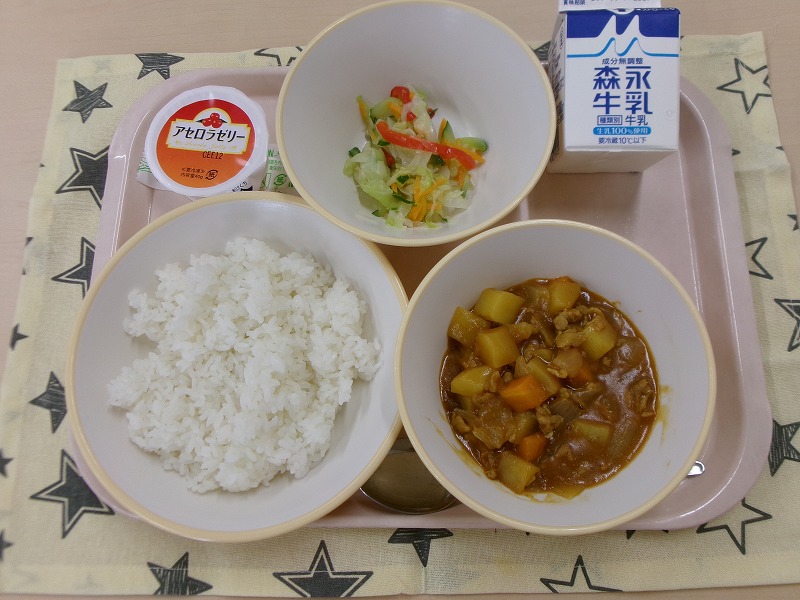 ピンクのトレーに給食が右上から反時計回りで牛乳、コールスローサラダ、アセロラゼリー、ご飯、カレーと並んでいる写真