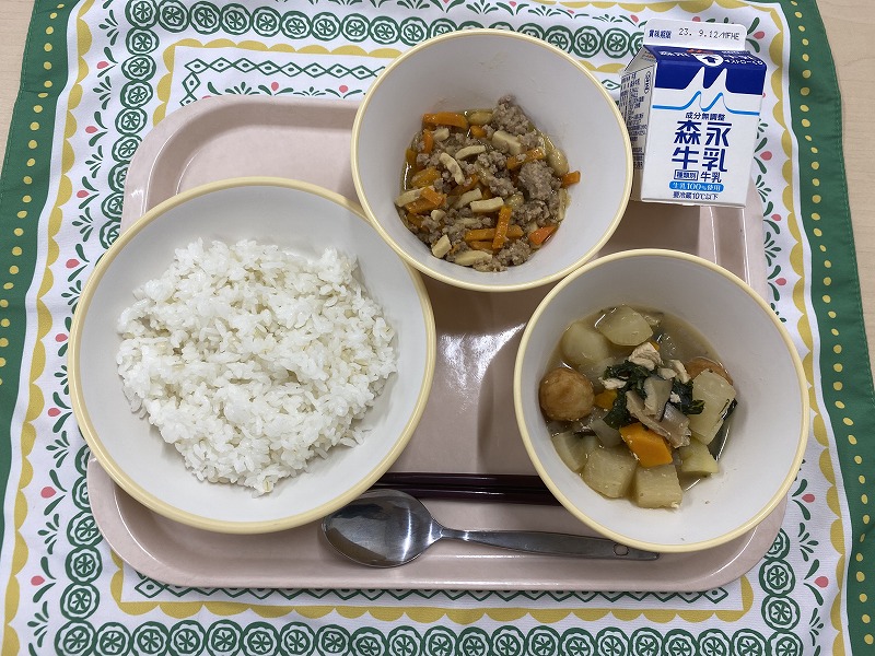 ピンクのトレーに給食が右上から反時計回りで牛乳、高野豆腐の肉そぼろ、もち麦入りごはん、和風ポトフと並んでいる写真