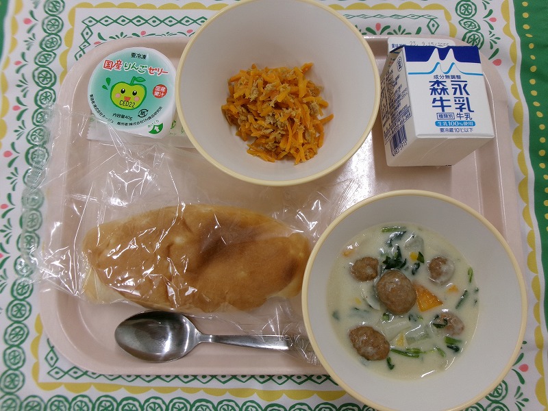 ピンクのトレーに給食が右上から反時計回りで牛乳、にんじんしりしり、りんごゼリー、玄米パン、ミートボールのクリームシチューと並んでいる写真