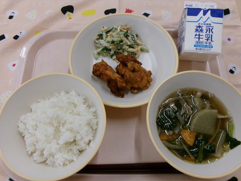 ピンクのトレーに給食が右上から反時計回りで牛乳、鶏肉のカレー揚げときゅうりのごまマヨ和え、ご飯、根菜汁と並んでいる写真