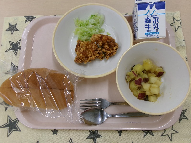 ピンクのトレーに給食が右上から反時計回りで牛乳、鶏の竜田揚げとキャベツ、パン、さつまいもとりんごの甘煮と並んでいる写真