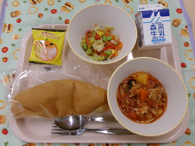 ピンクのトレーに給食が右上から反時計回りで牛乳、イタリアンサラダ、かぼちゃクリームもち、米粉コッペパン、豚肉のトマト煮と並んでいる写真