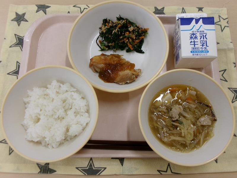 ピンクのトレーに給食が右上から反時計回りで牛乳、鶏肉の生姜焼きと小松菜とツナの和え物、ご飯、切り干し大根のピリ辛スープと並んでいる写真