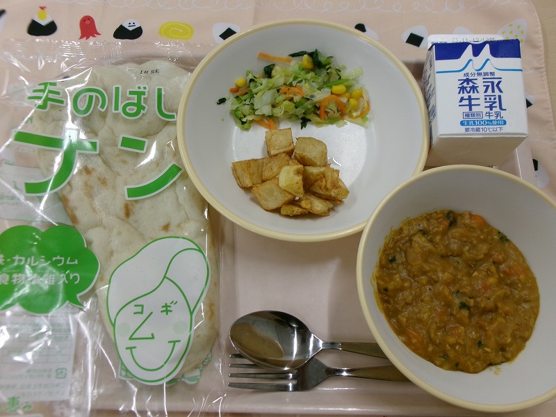 ピンクのトレーに給食が右上から反時計回りで牛乳、キャベツソテーとスパイシーポテト、ナン、ドライカレーと並んでいる写真
