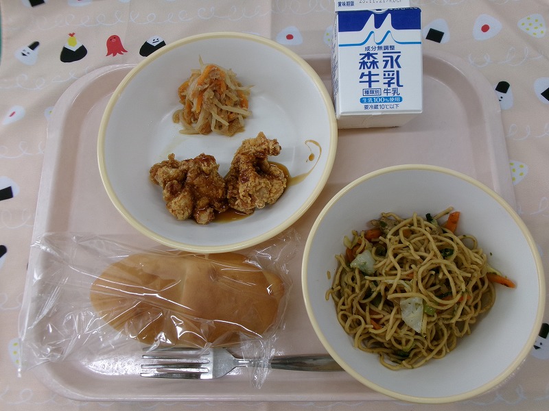 ピンクのトレーに給食が右上から反時計回りで牛乳、ツナサラダと照り焼きチキン、パン、ソース焼きそばと並んでいる写真