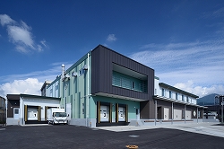 青い空の下、左奥に白いトラックが駐車され、複数のシャッター付きの搬入口がある秦野市学校給食センター外観を正面から広角で捉えた写真