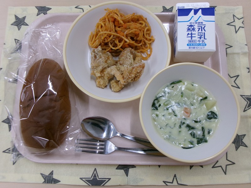 ピンクのトレーに給食が右上から反時計回りで牛乳、鶏のから揚げとスパゲティミートソース、黒パン、ほうれん草のミルクスープと並んでいる写真