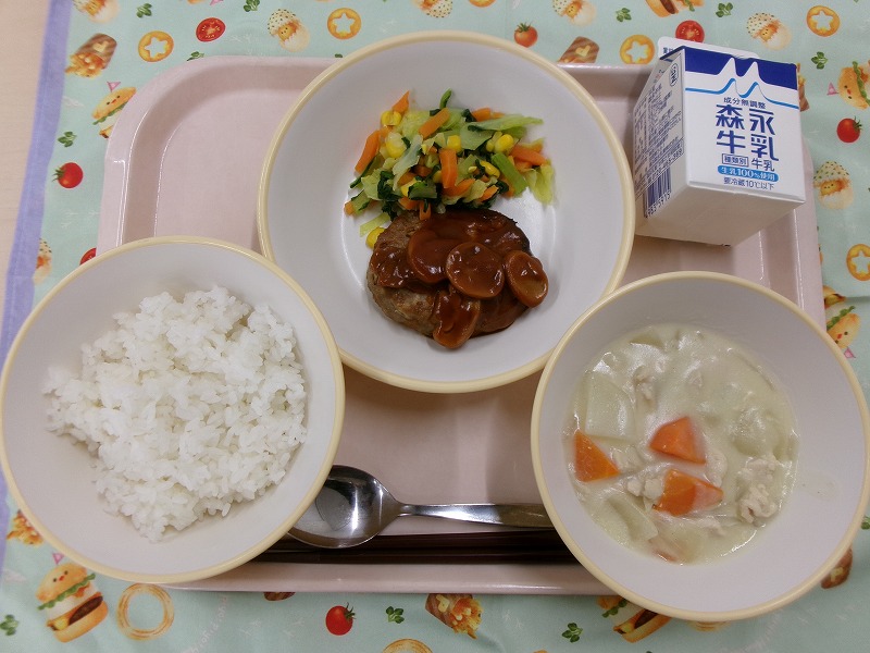 ピンクのトレーに給食が右上から反時計回りで牛乳、デミグラスハンバーグと野菜ソテー、ご飯、豆乳入りクリームスープと並んでいる写真