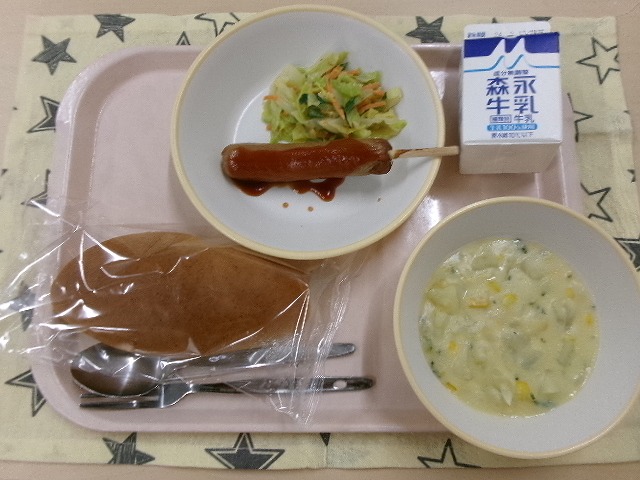 ピンクのトレーに給食が右上から反時計回りで牛乳、フランクフルトとごまドレッシングサラダ、秦野産小麦入りコッペパン、コーンシチューと並んでいる写真