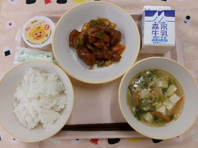 ピンクのトレーに給食が右上から反時計回りで牛乳、酢豚、いよかんゼリー、ご飯、五目スープと並んでいる写真