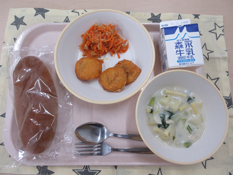 ピンクのトレーに給食が右上から反時計回りで牛乳、にんじんしりしりとチキンナゲット、黒パン、マカロニのクリーム煮と並んでいる写真