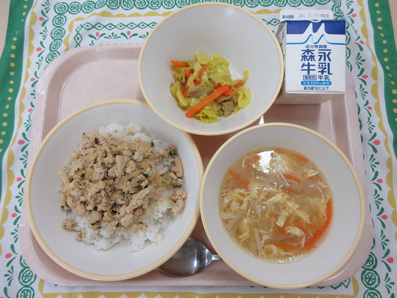 食が右上から反時計回りで牛乳、キャベツソテー、鶏そぼろ丼（麦入りごはん）、卵スープと並んでいる写真