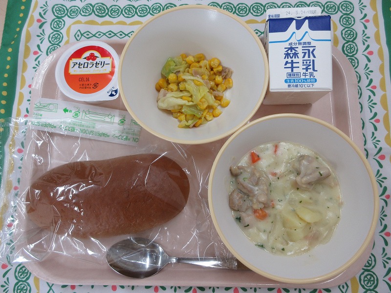 ピンクのトレーに給食が右上から反時計回りで牛乳、コーンとベーコンのソテー、アセロラゼリー、黒パン、チキンのクリームシチューと並んでいる写真