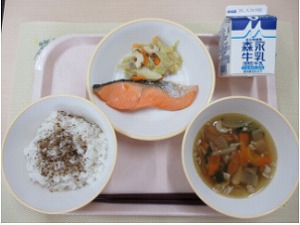 ピンクのトレーに給食が右上から反時計回りで牛乳、鮭の塩焼きと野菜のごま味噌炒め、ご飯（しそかつおふりかけ）、根菜汁と並んでいる写真