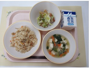 ピンクのトレーに給食が右上から反時計回りで牛乳、鶏肉とキャベツの旨塩炒め、かやくごはん、豆腐スープと並んでいる写真