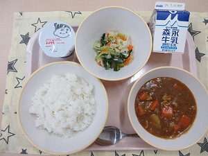 ピンクのトレーに給食が右上から反時計回りで牛乳、野菜ソテー、ヨーグルト、ご飯、カレーと並んでいる写真