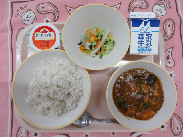 ピンクのトレーに給食が右上から反時計回りで牛乳、カラフルサラダ 、アセロラゼリー、麦入りごはん、夏野菜のカレーと並んでいる写真