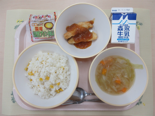 ピンクのトレーに給食が右上から反時計回りで牛乳、白身魚のバーベキューソース 、豆乳プリンタルト、コーンピラフ、コンソメスープと並べられている写真