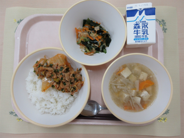 ピンクのトレーに給食が右上から反時計回りで牛乳、中華サラダ、豚キムチ丼（ごはん）、コーン中華スープと並べられている写真