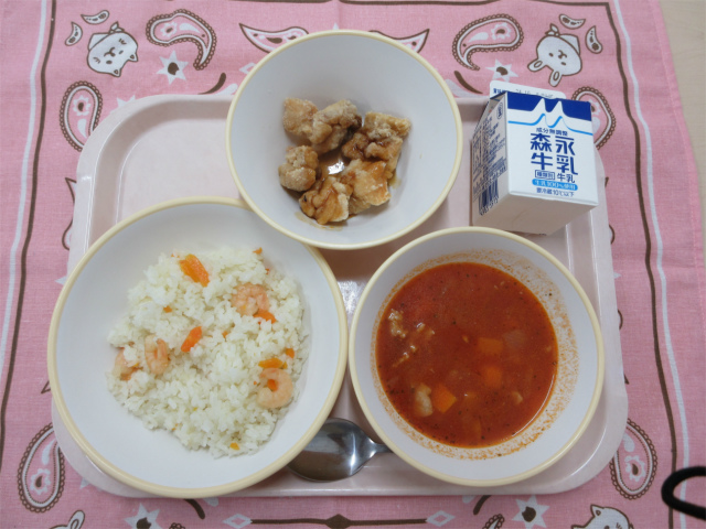 ピンクのトレーに給食が右上から反時計回りで牛乳、鶏肉の香味ソースがけ、えびピラフ、トマトスープと並べられている写真