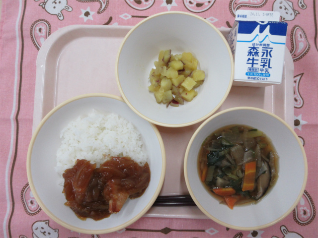 ピンクのトレーに給食が右上から反時計回りで牛乳、さつまいもの甘煮、ノギ屋の鶏飯（ごはん、鶏肉の生姜焼き）、けんちん汁と並べられている写真