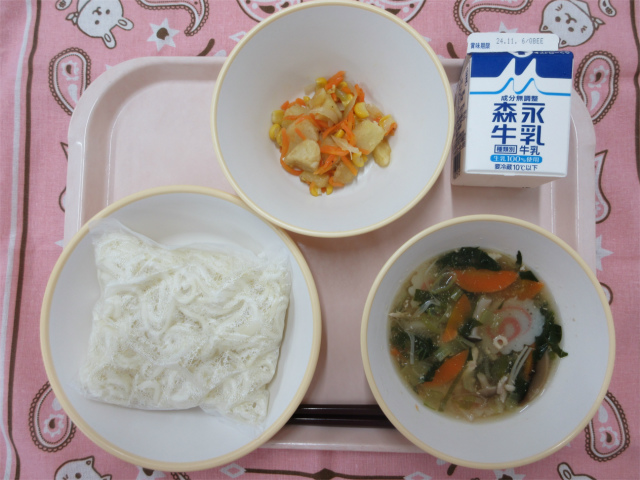 ピンクのトレーに給食が右上から反時計回りで牛乳、じゃがいものソテー、ソフト麺、きのこうどんのスープと並べられている写真