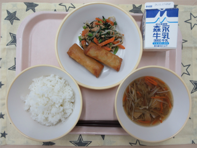 ピンクのトレーに給食が右上から反時計回りで牛乳、小松菜とツナの和え物、春巻き、ごはん、沢煮椀と並べられている写真