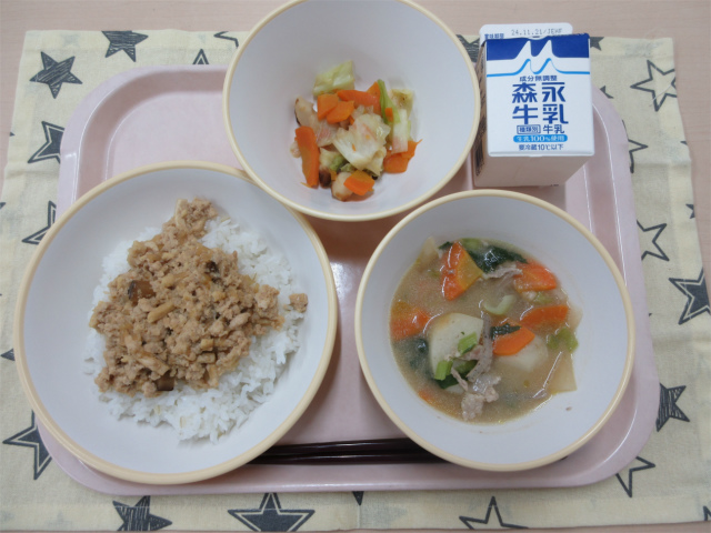 ピンクのトレーに給食が右上から反時計回りで牛乳、キャベツとちくわの炒め物、鶏そぼろ丼（もち麦入りご飯）、豚汁と並べられている写真